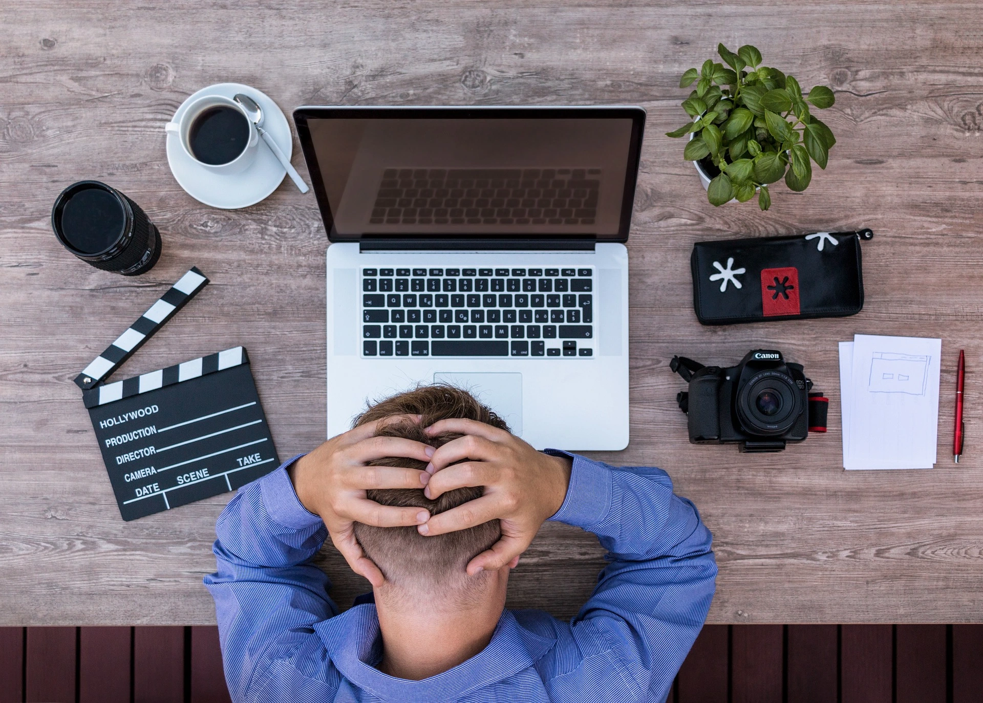Photo of a man in front of his laptop. He looks desperate and has his hands clasped over his head. On his table is a cup of coffee, a camera lens, a film flap, a pencil case, some paper and a camera. StudySmarter Magazine