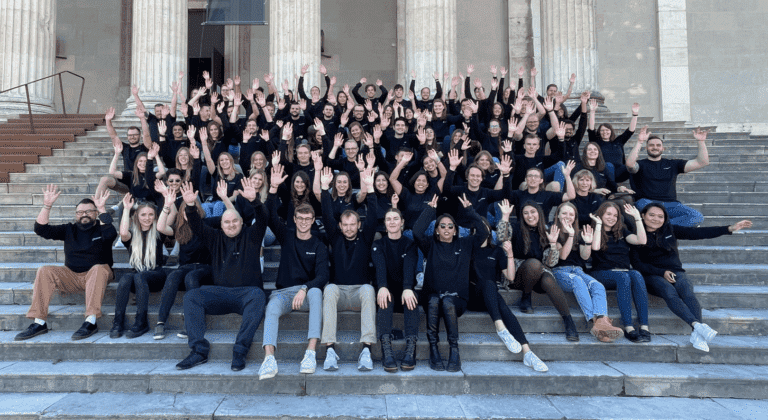 A group of people from the Vaia office sitting at the steps of a building and cheering to welcome people starting their careers. Vaia Magazine