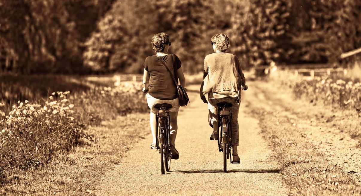 Two women riding bicycles down a woodland park lane as a quick study break exercise. Vaia Magazine
