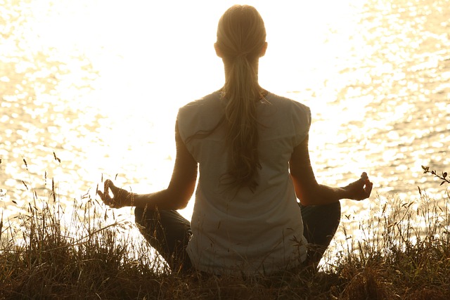 A woman with her back to the camera and sitting cross legged and looking at a lake at sunset while meditating. Vaia Magazine