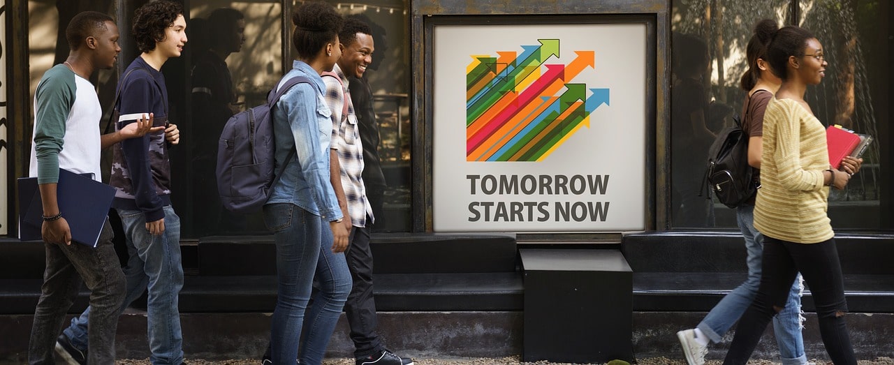A group of students walking past a motivational poster saying 'Tomorrow Starts Now'. Vaia Magazine